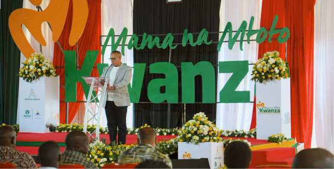 Speaker at a lectern in front of large Mama na Mtoto Kwanza stage letters and flower displays.