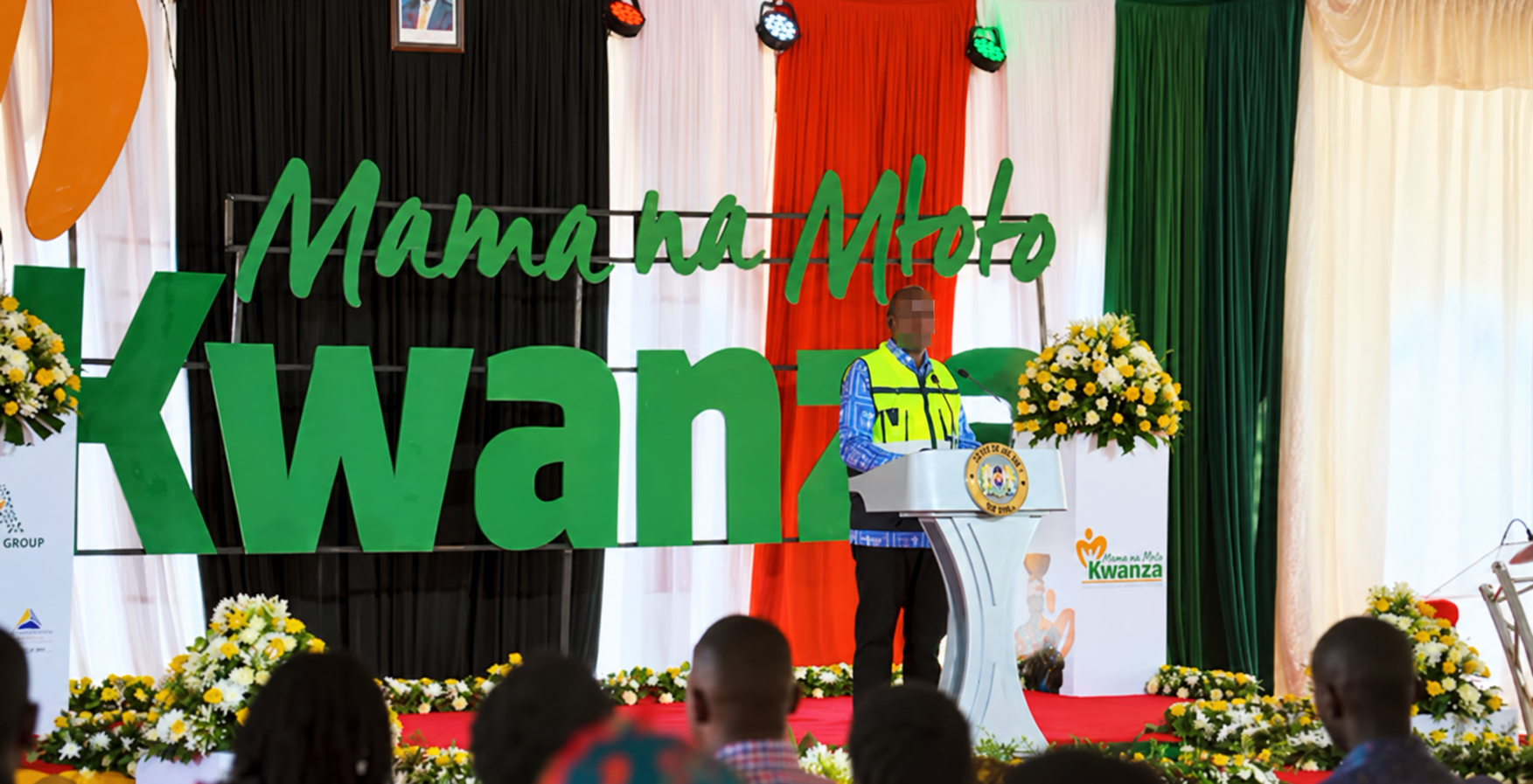 Speaker at a podium with large green Mama na Mtoto Kwanza stage letters behind him.
