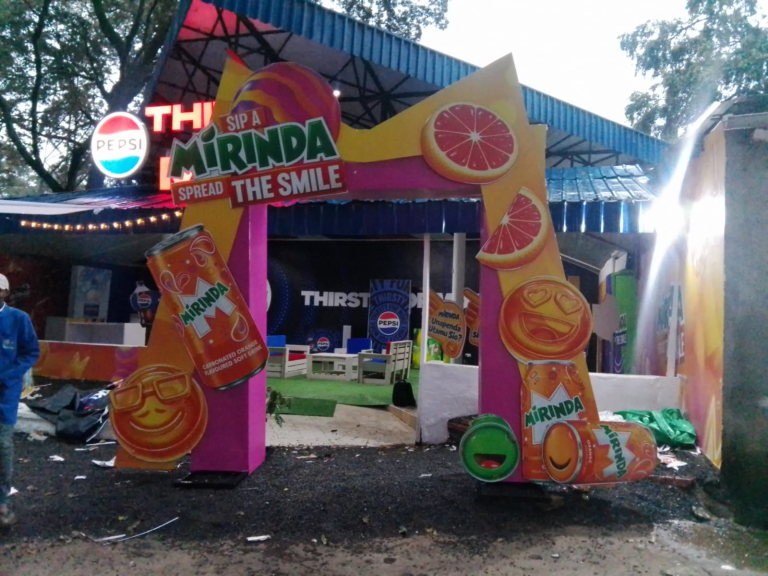 Pink and yellow Mirinda entrance arch with drink can and fruit graphics at an outdoor venue.